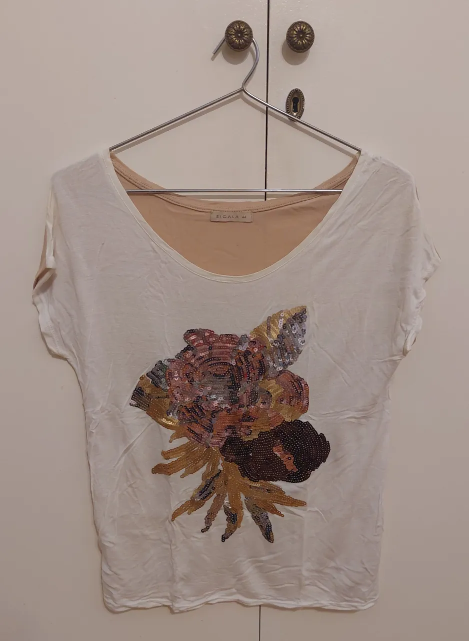 Remera con frente color crudo y  estampado floral de lentejuelas en tonos dorados, rosas y marrones. La espalda es color beige. Tiene una pequña mancha en la espalda, d3l laso izquierdo. Ideal para darle un toque chic a tu outfit.