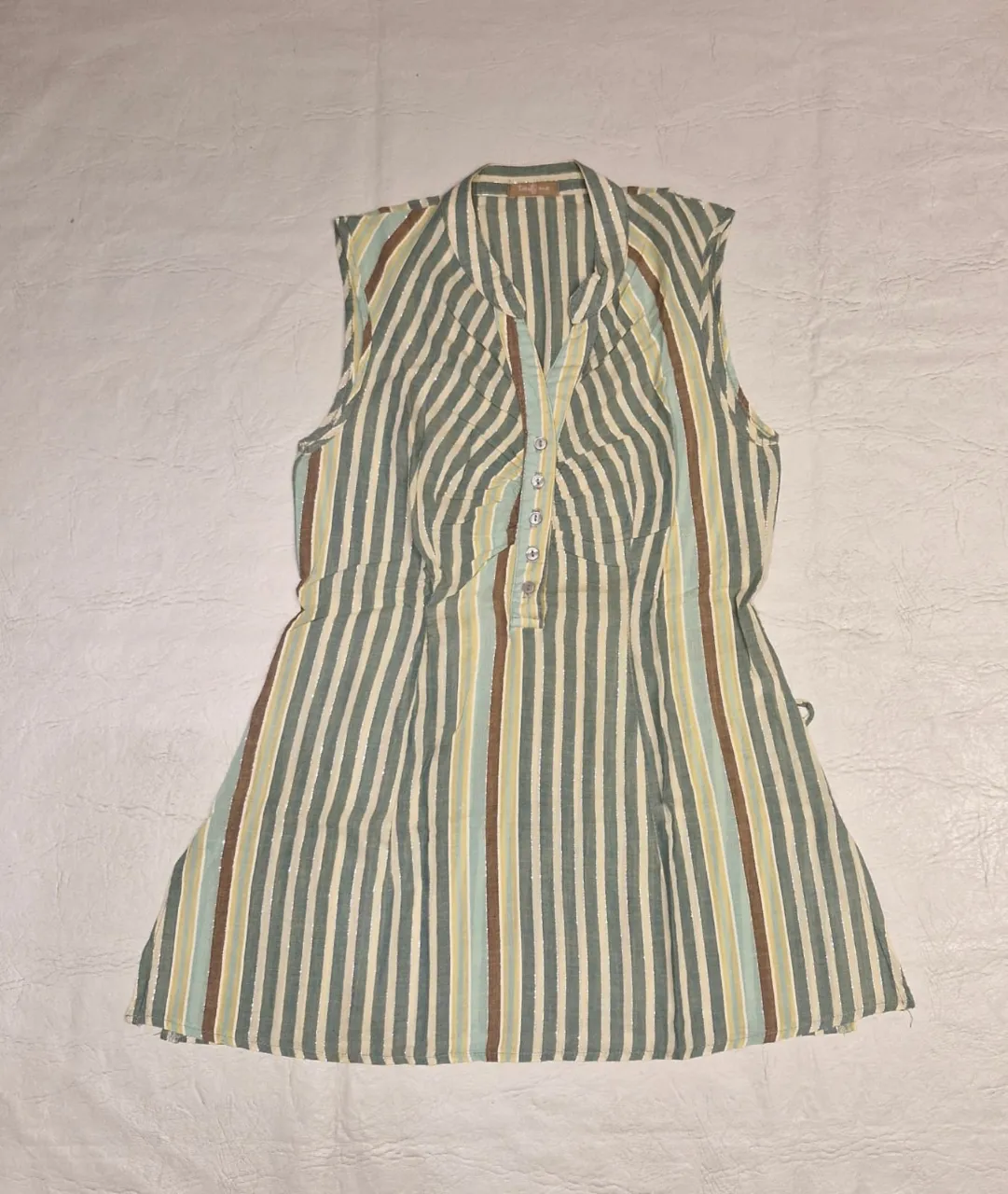Blusa sin mangas a rayas verticales en tonos verde, amarillo y marrón, con hilos dorados. Cuello camisero y botones frontales. Ideal para un look fresco y casual. Con dos pequeñas trabas en los costados para pasar un lazo (no incluido)