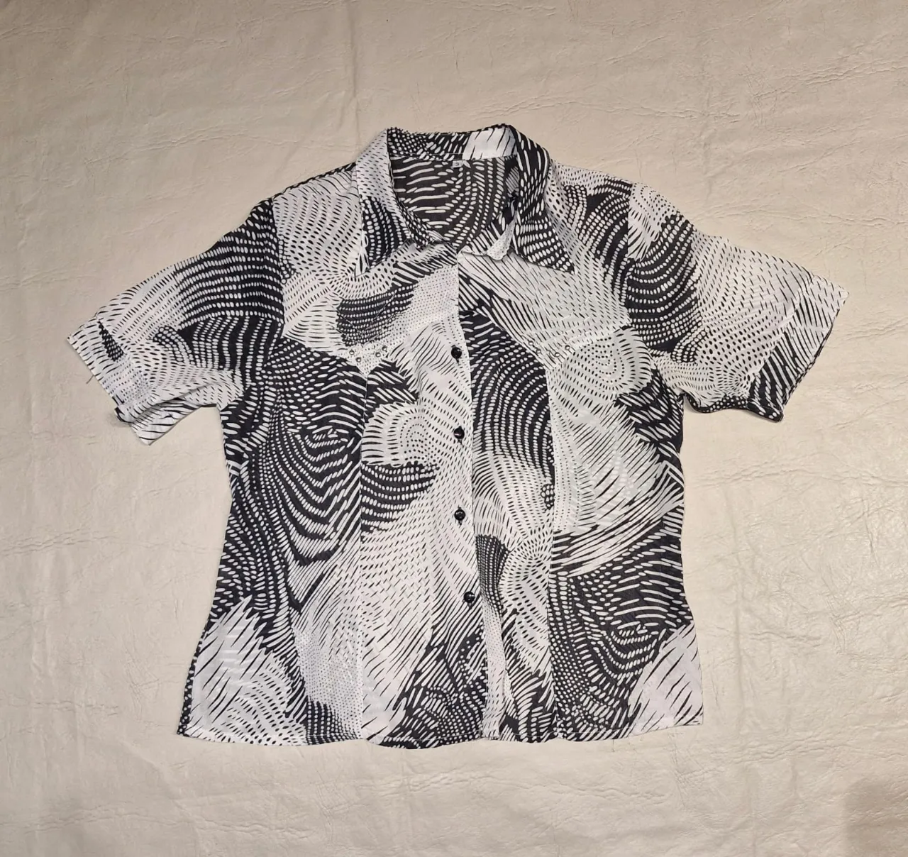 Camisa estampada con diseño abstracto en blanco y negro. Corte clásico, ideal para un look casual o semi-formal. Con hombreras pequeñas, forradas en la misma tela estampada. Con un diseño bordado y tachas plateadas en el frente. Perfecta para combinar con jeans o pantalones de vestir.