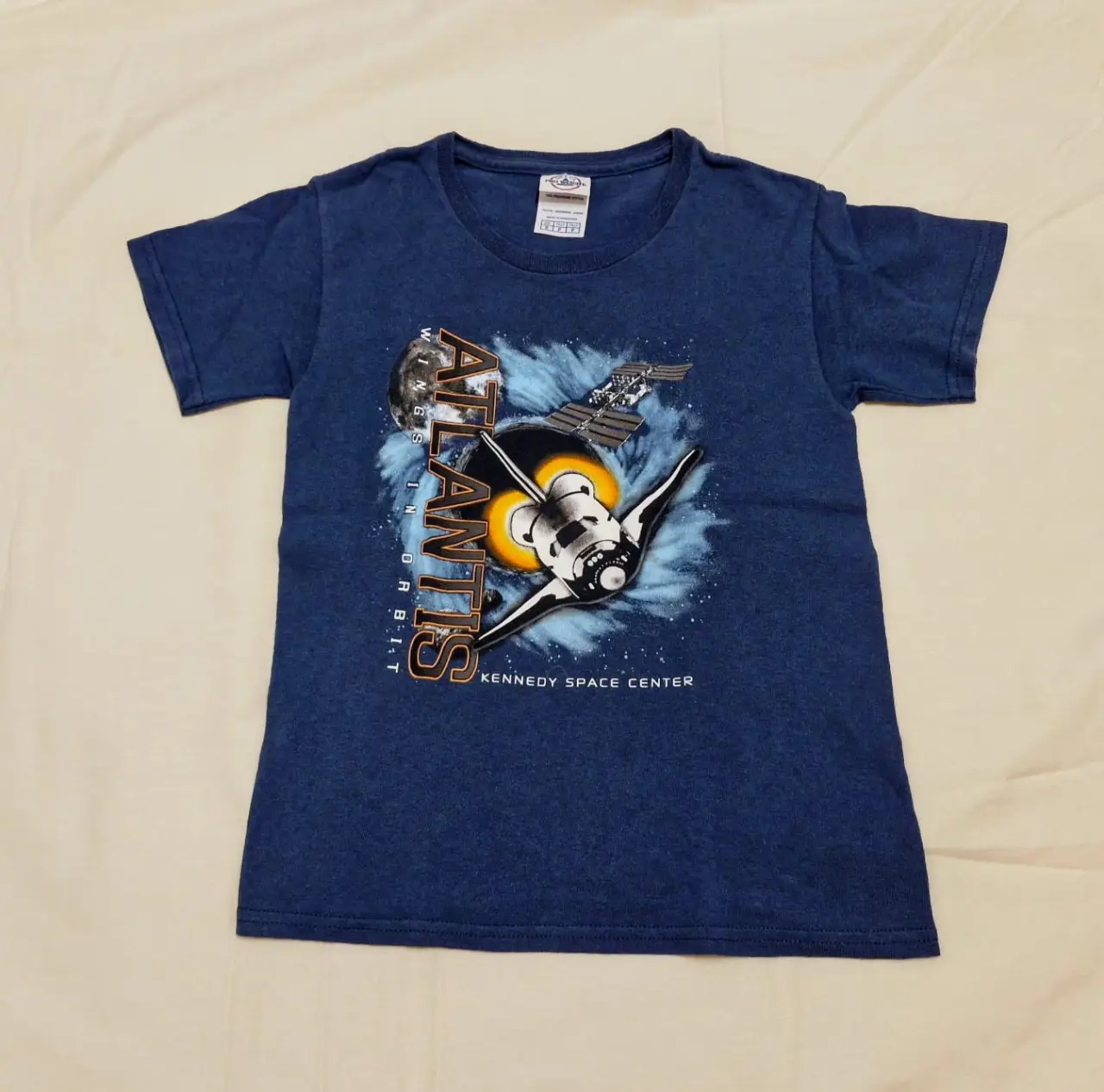 Remera azul de manga corta para niños con estampado del transbordador espacial Atlantis y el Kennedy Space Center. Ideal para fanáticos del espacio y la ciencia.