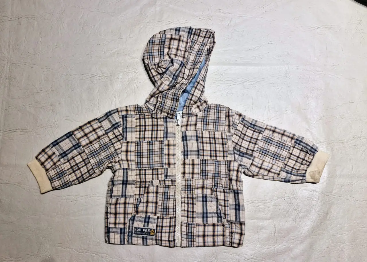 Campera liviana con capucha para niño, con diseño a cuadros en tonos marrones, azules y blancos. Cuenta con cierre frontal y una etiqueta pequeña del lado inferior derecho con la inscripción "hot dog". Ideal para un look casual y abrigado.