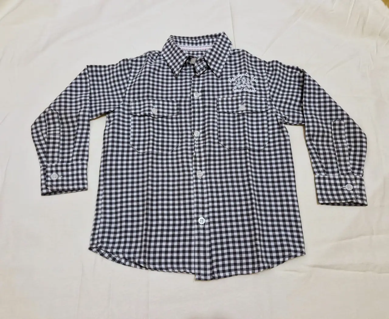 Camisa a cuadros gris y blanco para niño, manga larga, con dos bolsillos delanteros y detalles bordados. Ideal para un look casual y cómodo.