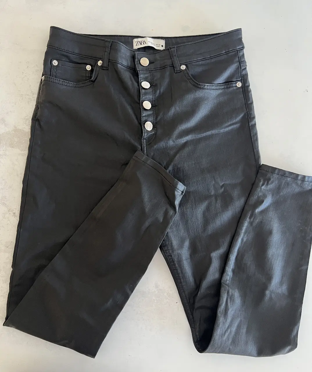 Pantalón de vestir negro de cuero sintético de la marca Zara. Tiro alto, con botones plateados y bolsillos delanteros. Ideal para un look moderno y elegante.
