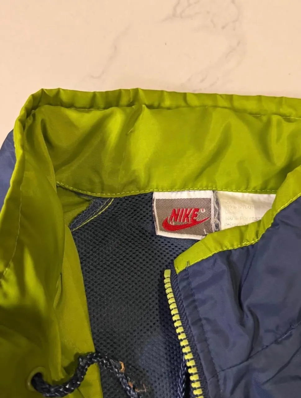 Campera deportiva Nike azul con paneles laterales y mangas en verde lima. Cierre frontal amarillo y cordón ajustable en la cintura.
