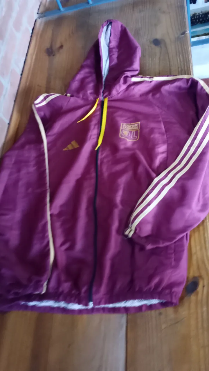 Buzo con cierre y capucha, color violeta con detalles amarillos. Con logo del equipo Olympique Lyonnais y logo de Adidas. Ideal para usar en días frescos.