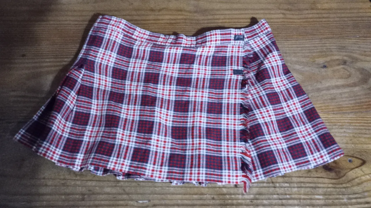Pollera mini escocesa para mujer, con diseño a cuadros en rojo y blanco. Presenta detalles de botones y un corte asimétrico que le da un toque moderno. Ideal para un look casual y con estilo.
