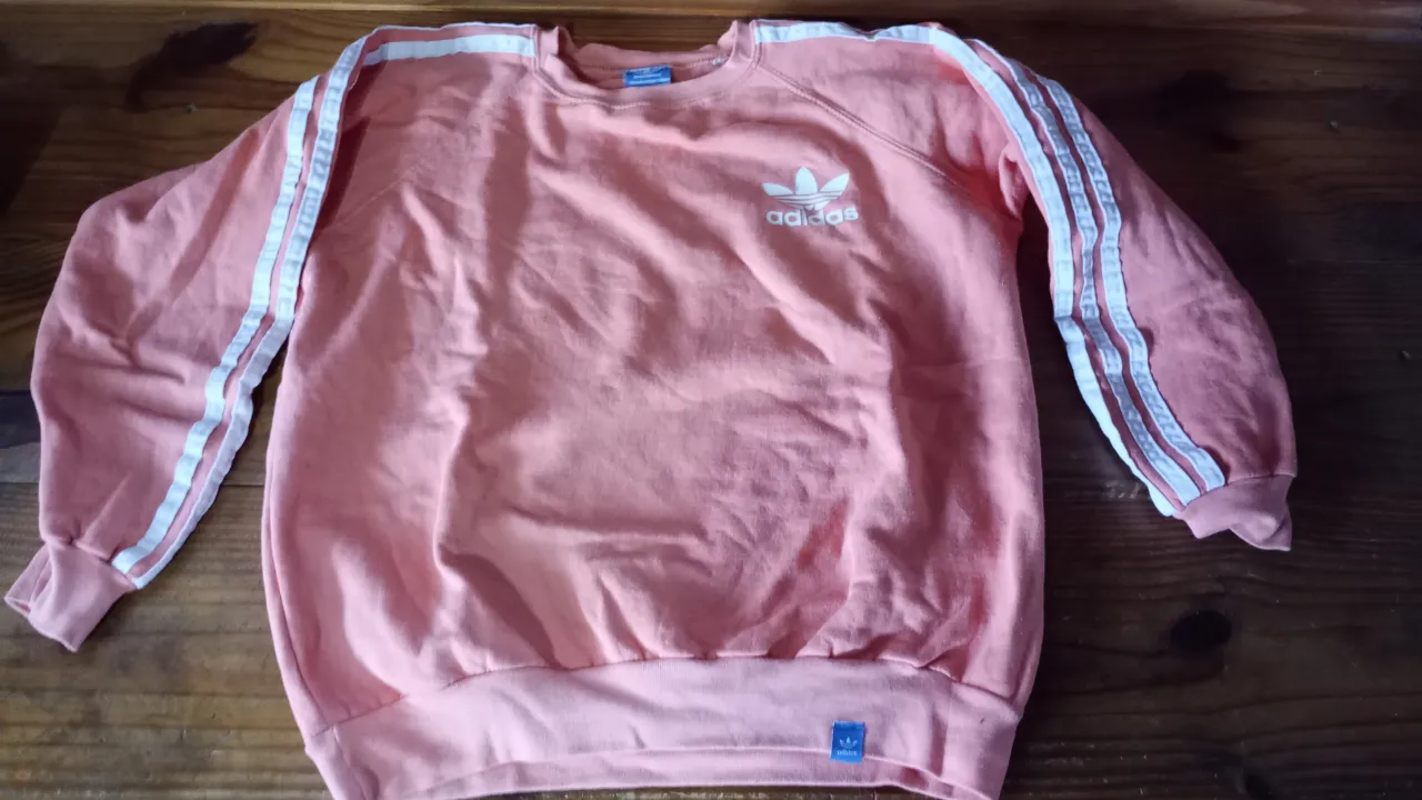 Buzo Adidas de cuello redondo en color rosa, con las clásicas tres tiras blancas en las mangas y el logo de la marca en el pecho. Ideal para un look deportivo y casual.