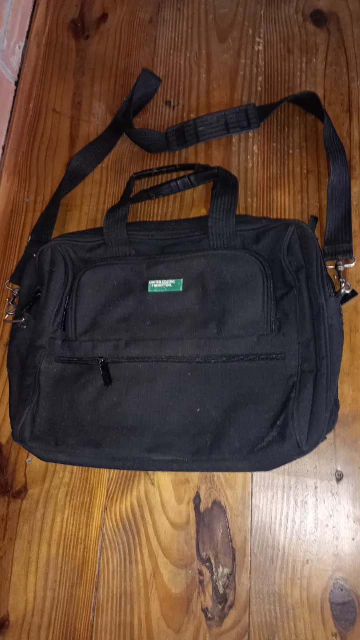 Bolso de viaje/porta notebok negro de la marca United Colors of Benetton, con correa ajustable y manijas superiores. Cuenta con un bolsillo frontal con cierre y un compartimento principal espacioso. Ideal para viajes cortos o como equipaje de mano.