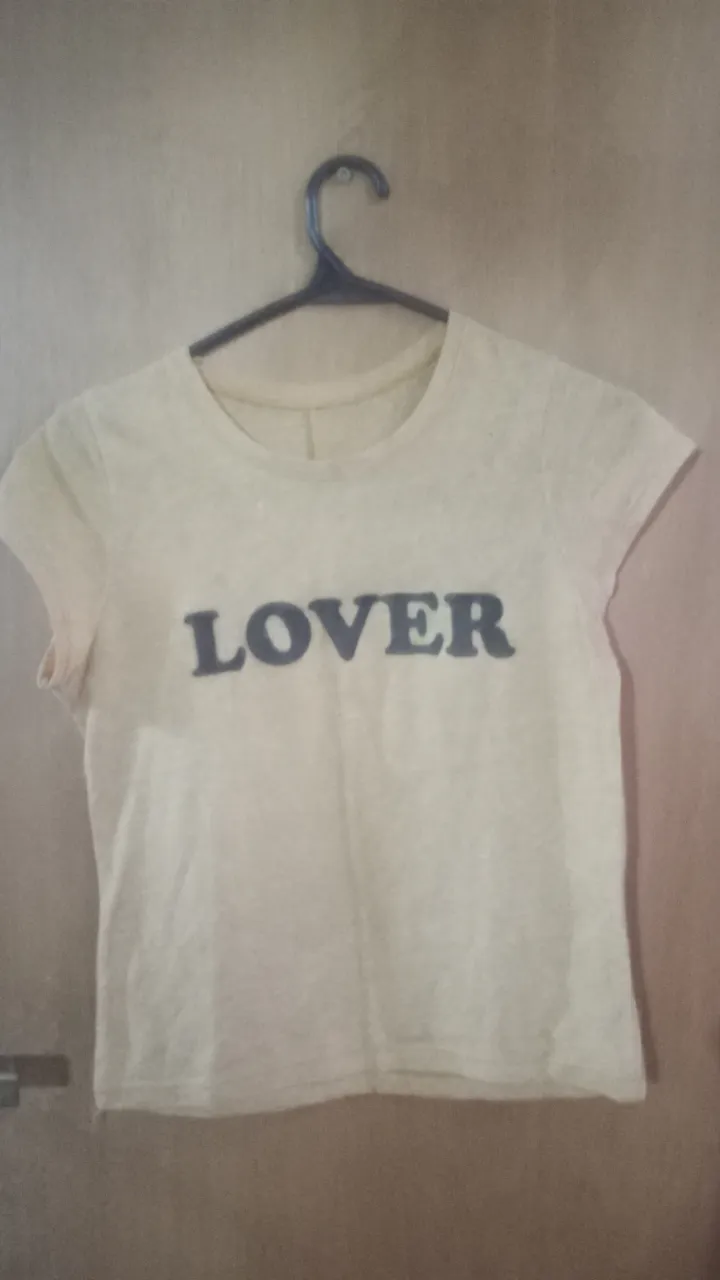 Remera de mujer de color amarillo claro con la palabra 'LOVER' estampada en negro en el frente. Tiene un bordado de una rosa roja en la manga. Ideal para un look casual y con onda.