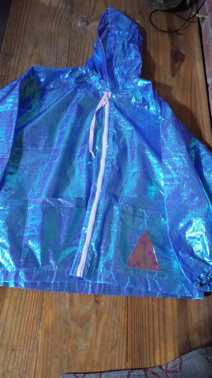 Rompevientos impermeable azul tornasolado con capucha y cierre rosa. Posee bolsillos delanteros y un detalle triangular en un bolsillo. Ideal para la lluvia o recitales o un look moderno.