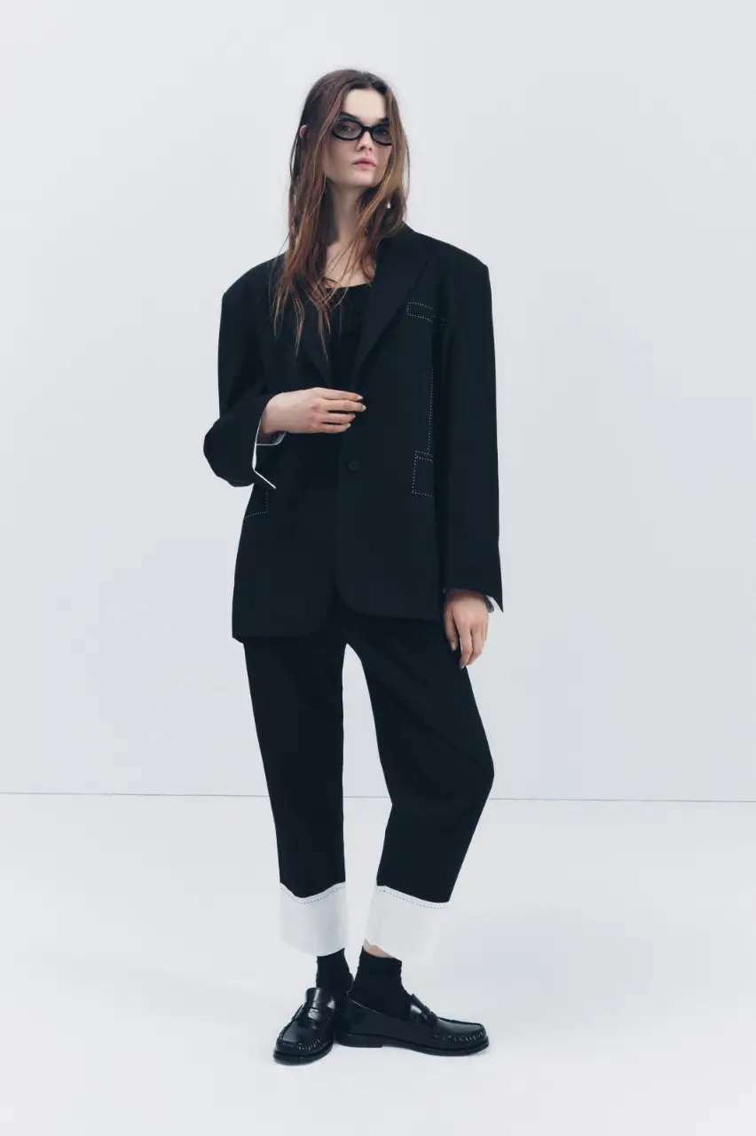 Pantalón sastrero negro con detalle de popelín blanco en el bajo y pespuntes. Tiro medio con trabillas, bolsillos laterales y falsos bolsillos en la espalda. Cierre frontal con cremallera, botón interior y ganchos metálicos. Un clásico renovado para un look elegante y moderno.