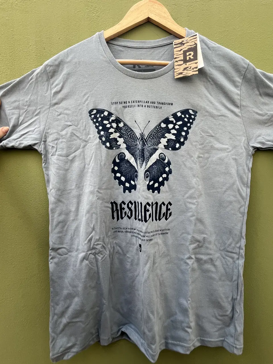 Remera Rever gris azulada con estampado de mariposa y la palabra 'Resilience'. Cuello redondo y manga corta. 100% algodón, nueva sin uso. 