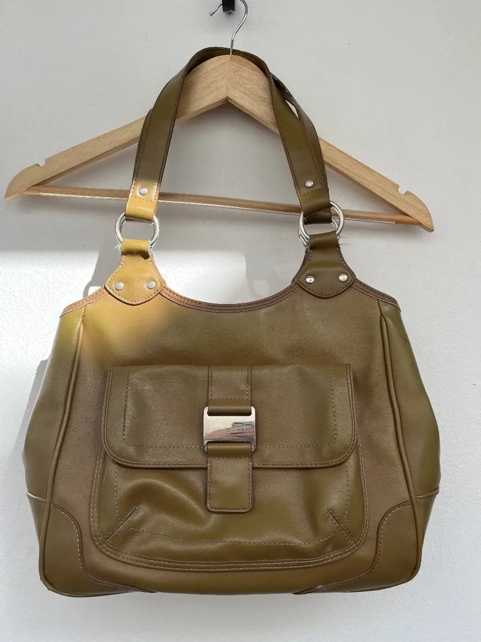 Cartera Nine West de cuero sintético en tono marrón verdoso. Cuenta con un bolsillo frontal con cierre de hebilla y un amplio compartimento interior. De afuera se ve nueva, por dentro tiene algunos detalles.