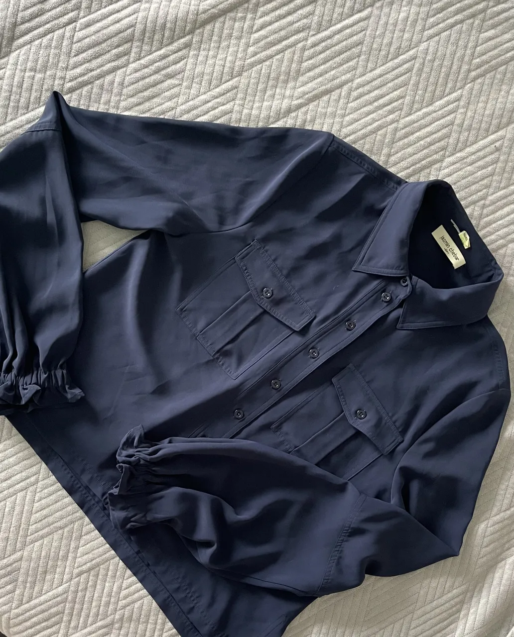 Blusa de mujer azul marino de la marca Jazmin Chebar, con mangas largas terminadas en puños elastizados. Posee dos bolsillos delanteros y botones a tono. Ideal para un look elegante y versátil.