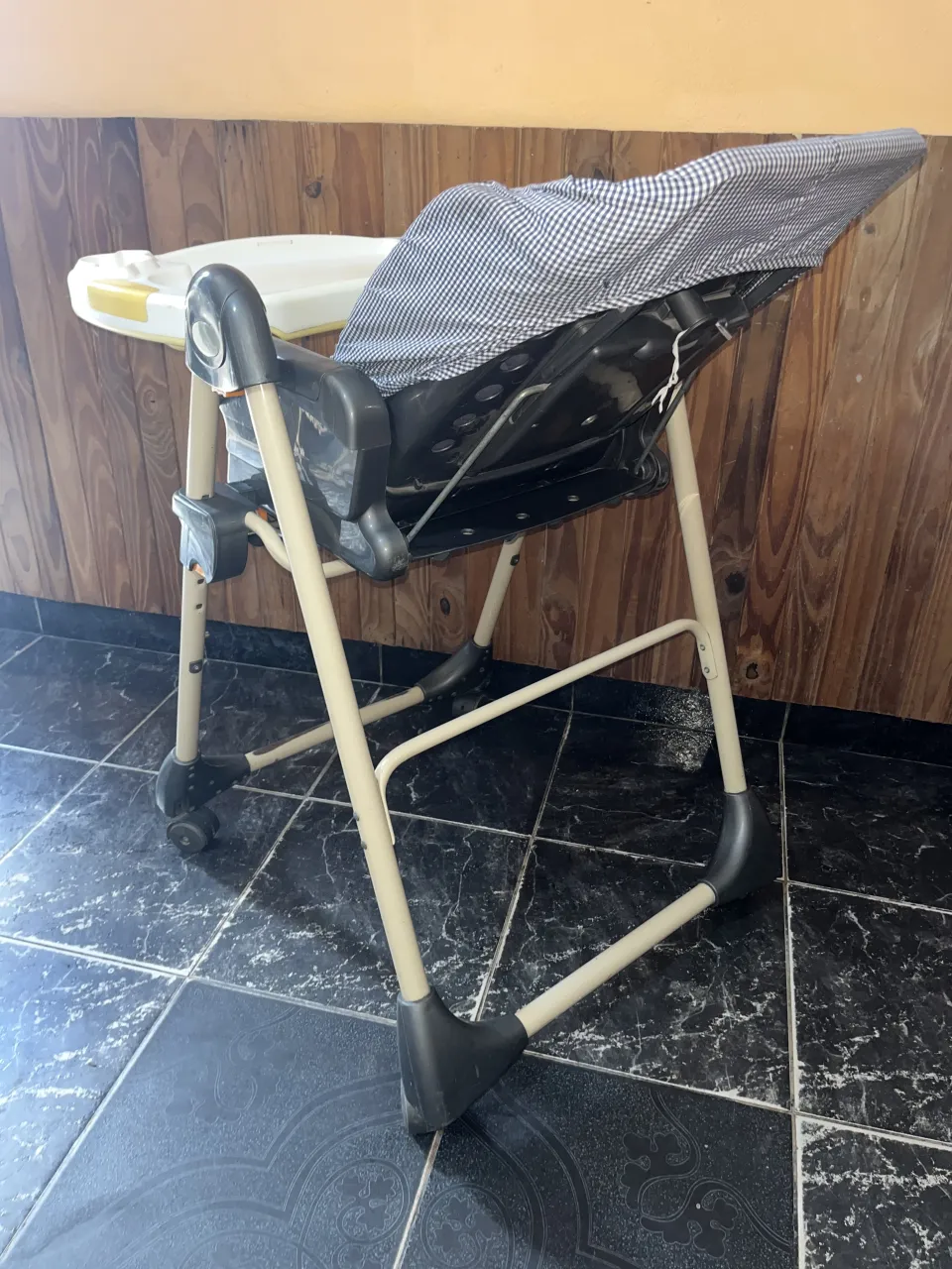 Silla de comer para bebé, plegable, con bandeja desmontable y funda acolchada a cuadros. Estructura metálica color beige y detalles en negro. Ruedas en las patas delanteras y trasera. 
