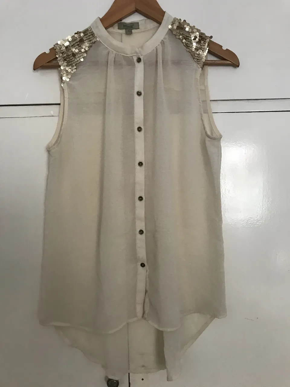 Blusa sin mangas de color crudo transparente, con detalles de lentejuelas doradas en los hombros. Tiene botones en la parte delantera y un diseño elegante. Ideal para un look sofisticado.