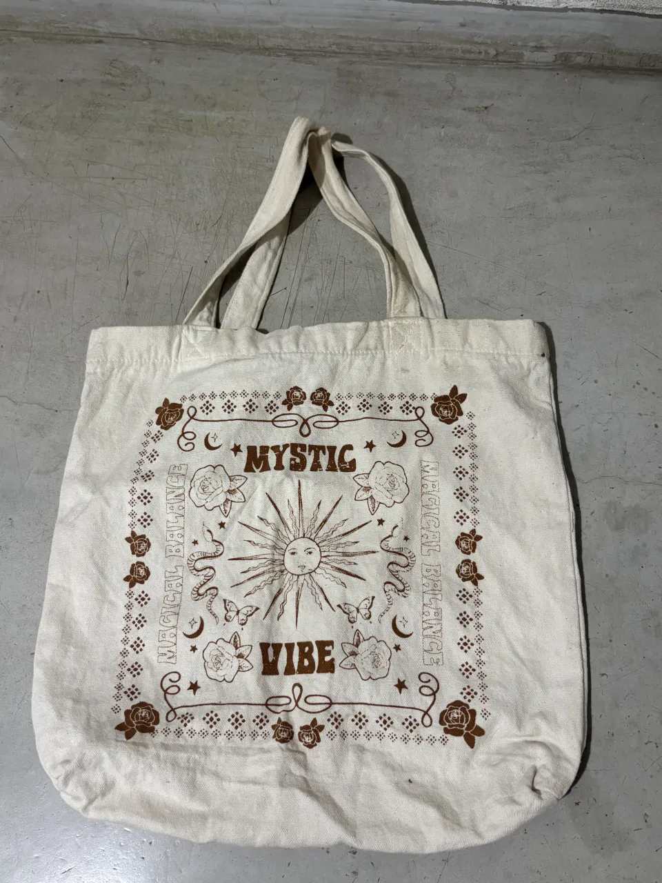 Bolso tote de tela color crudo con estampa "Mystic Vibe" en marrón. Diseño con sol, estrellas, mariposas y flores. Ideal para el día a día, para llevar tus esenciales con un toque místico.