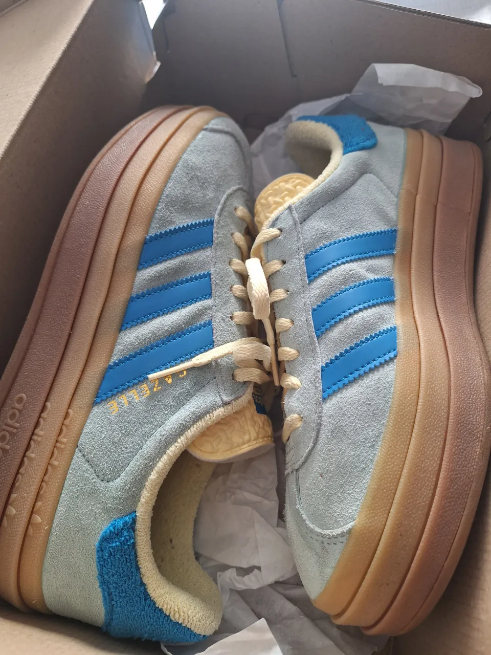 Zapatillas Adidas Gazelle con plataforma. Color gris claro con detalles en azul y cordones amarillos.