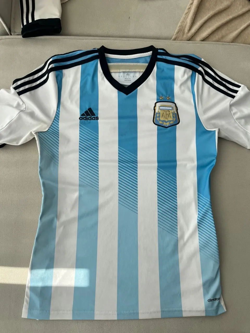 Remera de fútbol a rayas verticales celestes y blancas. Escudo de la AFA y logo de Adidas en el frente. Tiras negras con detalles blancos en los hombros.