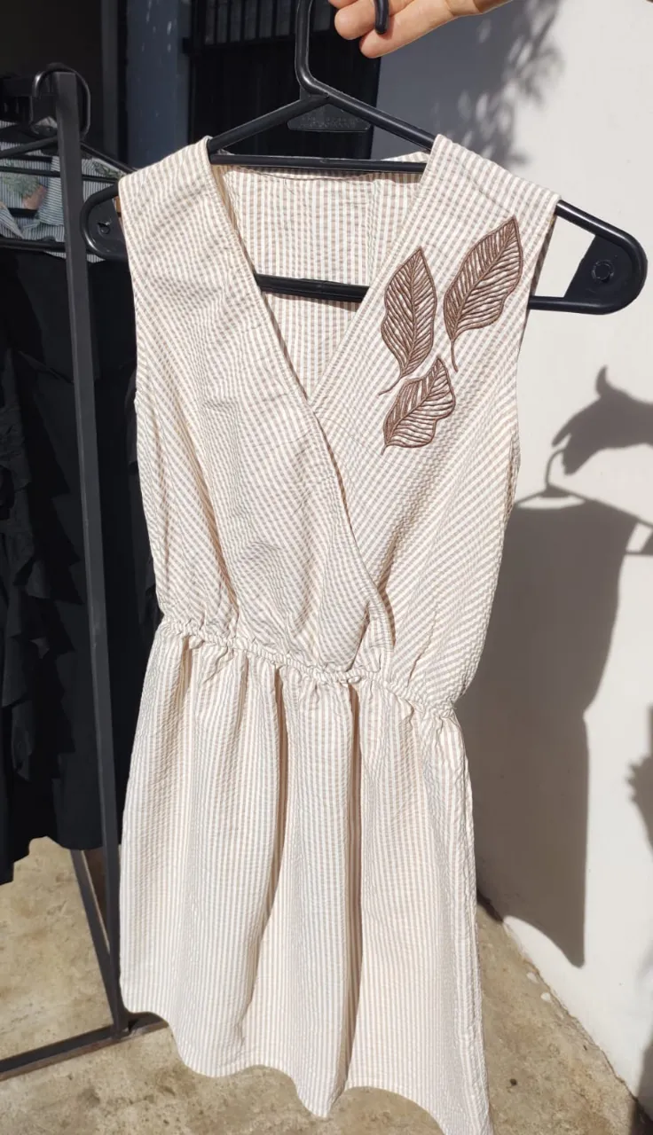 Vestido corto de tela rayada en blanco y beige. Posee un cuello en V cruzado y elástico en la cintura. Tiene un bordado de hojas en color marrón en la parte superior izquierda.