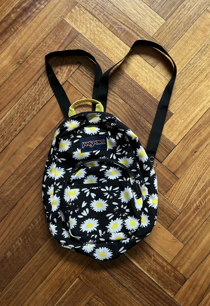 Mochila JanSport con estampado de flores blancas sobre fondo negro. Ideal para uso diario o para llevar tus cosas con estilo.