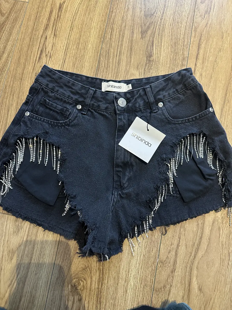 Short de jean negro con detalles de flecos de strass y bolsillos. Ideal para un look de fiesta o salida.