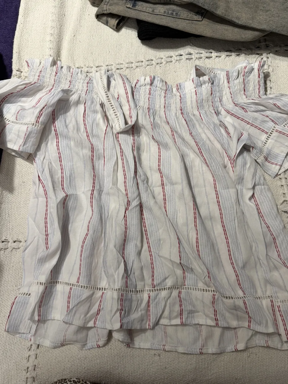 Blusa blanca con rayas finas rojas y azules, escote con elástico y volados, mangas cortas con volados y detalles de puntilla. Ideal para un look fresco y casual de día.