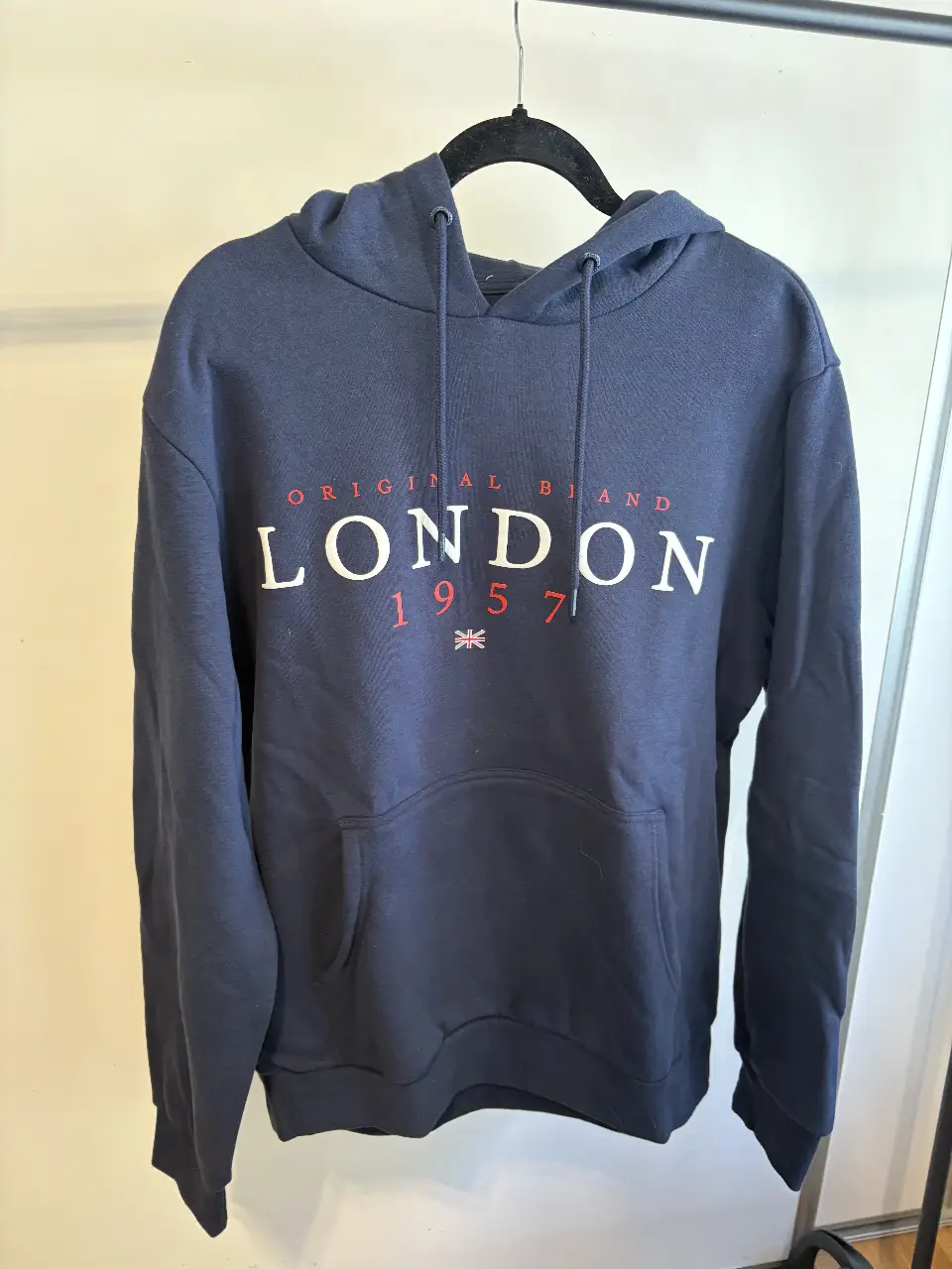 Buzo canguro de hombre, color azul marino, con estampado "LONDON 1957" en blanco y rojo. Tiene capucha con cordones y bolsillo frontal tipo canguro. Talle L.