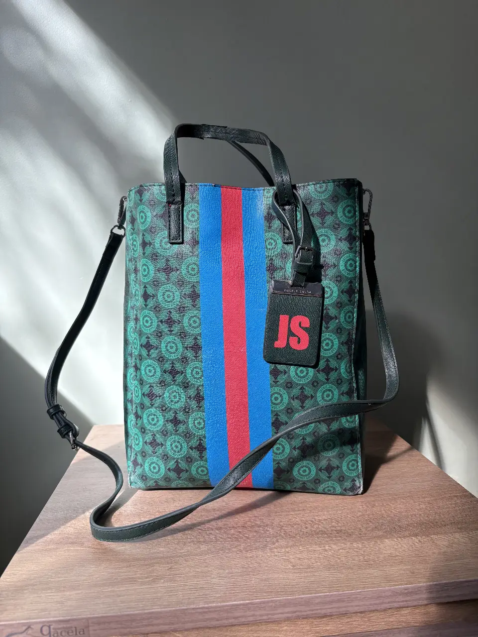 Bolso tote de diseño con estampado geométrico en tonos verdes, con franja central en rojo y azul. Incluye correa ajustable y etiqueta con iniciales. Ideal para un look casual y moderno.