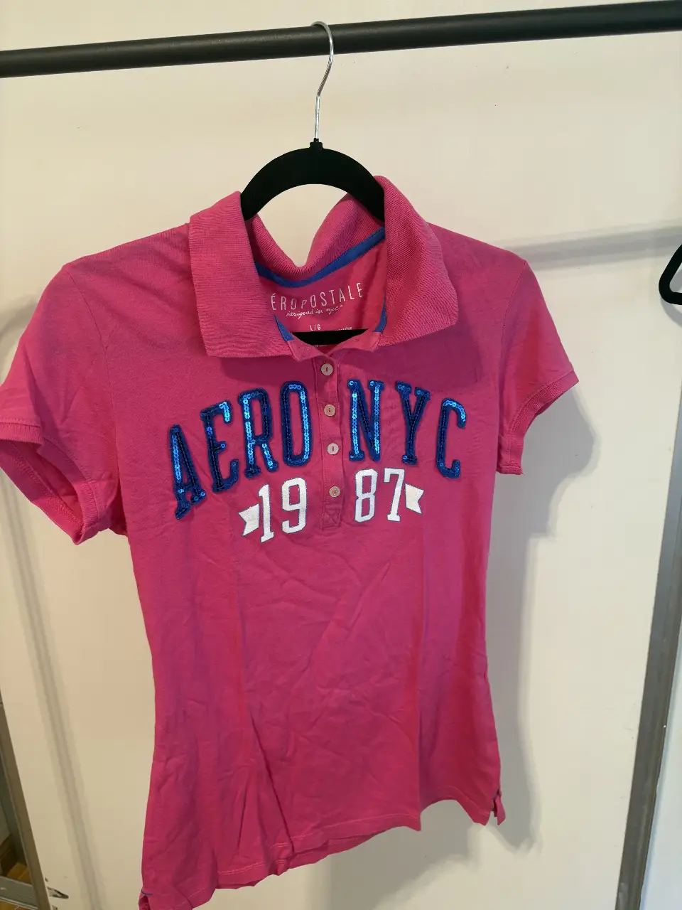 Remera rosa de mujer, estilo polo, con detalles en azul y el logo "AERO NYC 1987". Ideal para un look casual y deportivo.