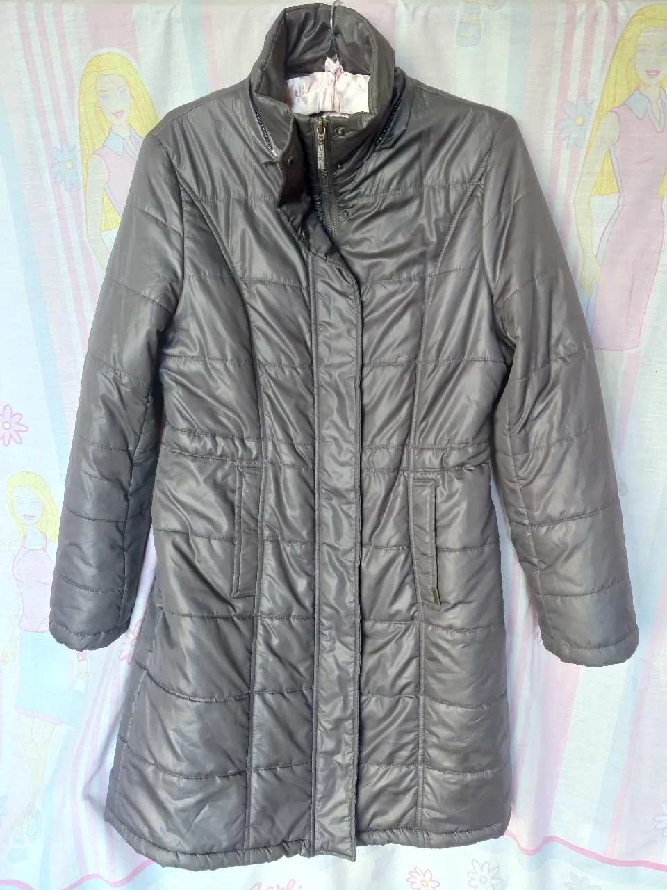 Campera larga acolchada de color gris, ideal para el invierno. Cuenta con cierre y bolsillos. Tiene un detalle en la cintura para ajustar.