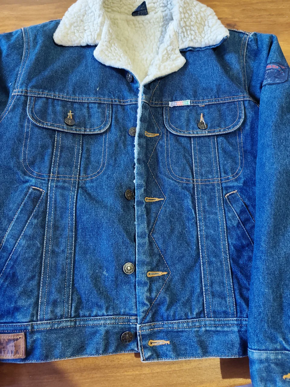 Campera de jean azul con cuello de corderito blanco. Posee dos bolsillos superiores con tapa y botones metálicos. Cierre de botones metálicos en el frente. Tiene parches en las mangas y etiquetas de la marca "Montana".