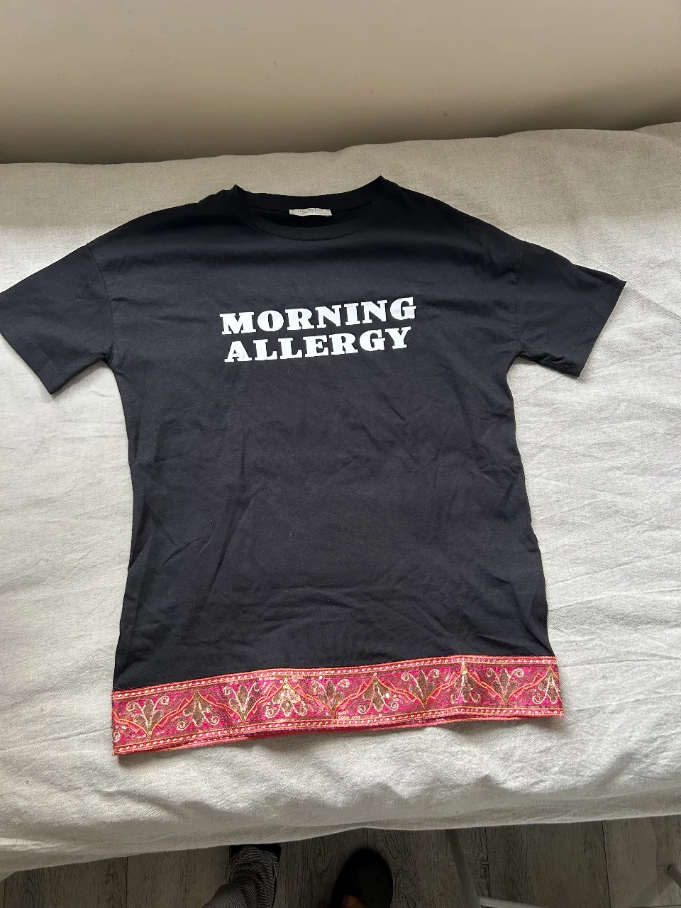 Remera negra de algodón con estampado "MORNING ALLERGY" en blanco. Detalles de bordado rosa y dorado en el borde inferior. Ideal para un look casual y con onda.