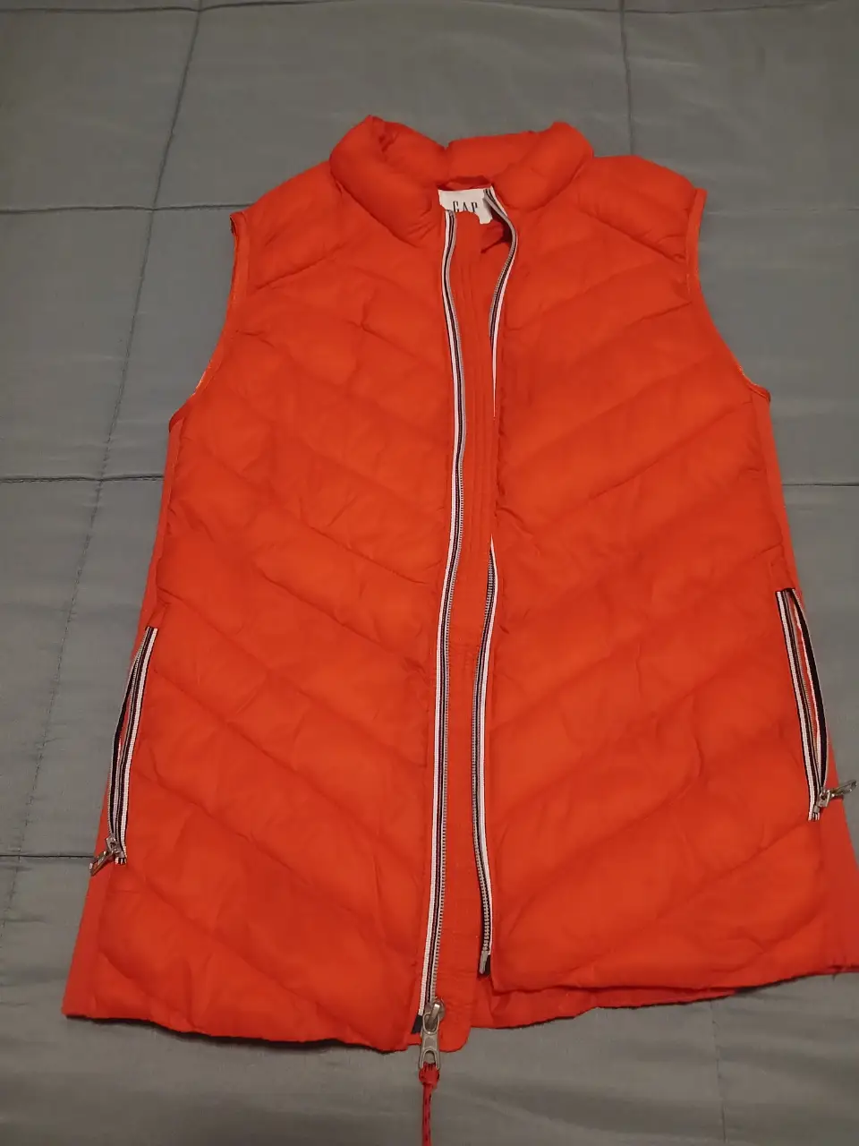 Chaleco acolchado naranja de la marca GAP, con cierre y bolsillos con detalles en blanco y negro. Ideal para un look casual y abrigado.