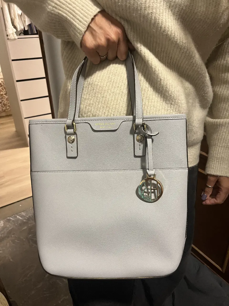 Bolso tote de color celeste, con manijas y correa desmontable. Cuenta con un diseño elegante y funcional, ideal para llevar tus pertenencias con estilo. Incluye un compartimento interno con cierre y detalles dorados.