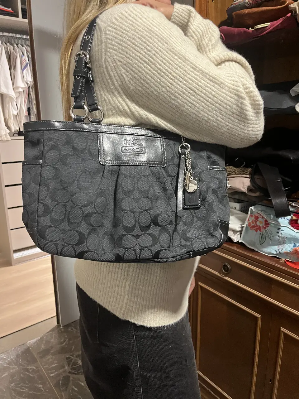 Cartera Coach negra con monograma, ideal para llevar al hombro. Cuenta con detalles de cuero y un dije con el logo de la marca. Perfecta para un look casual y elegante.