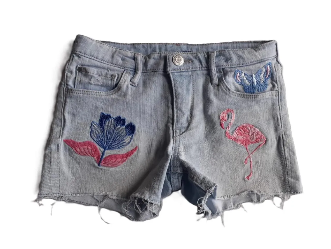 Short de jean celeste claro para niña, con detalles bordados de flores y un flamenco. Tiene un corte desflecado y un talle 8-9 años.