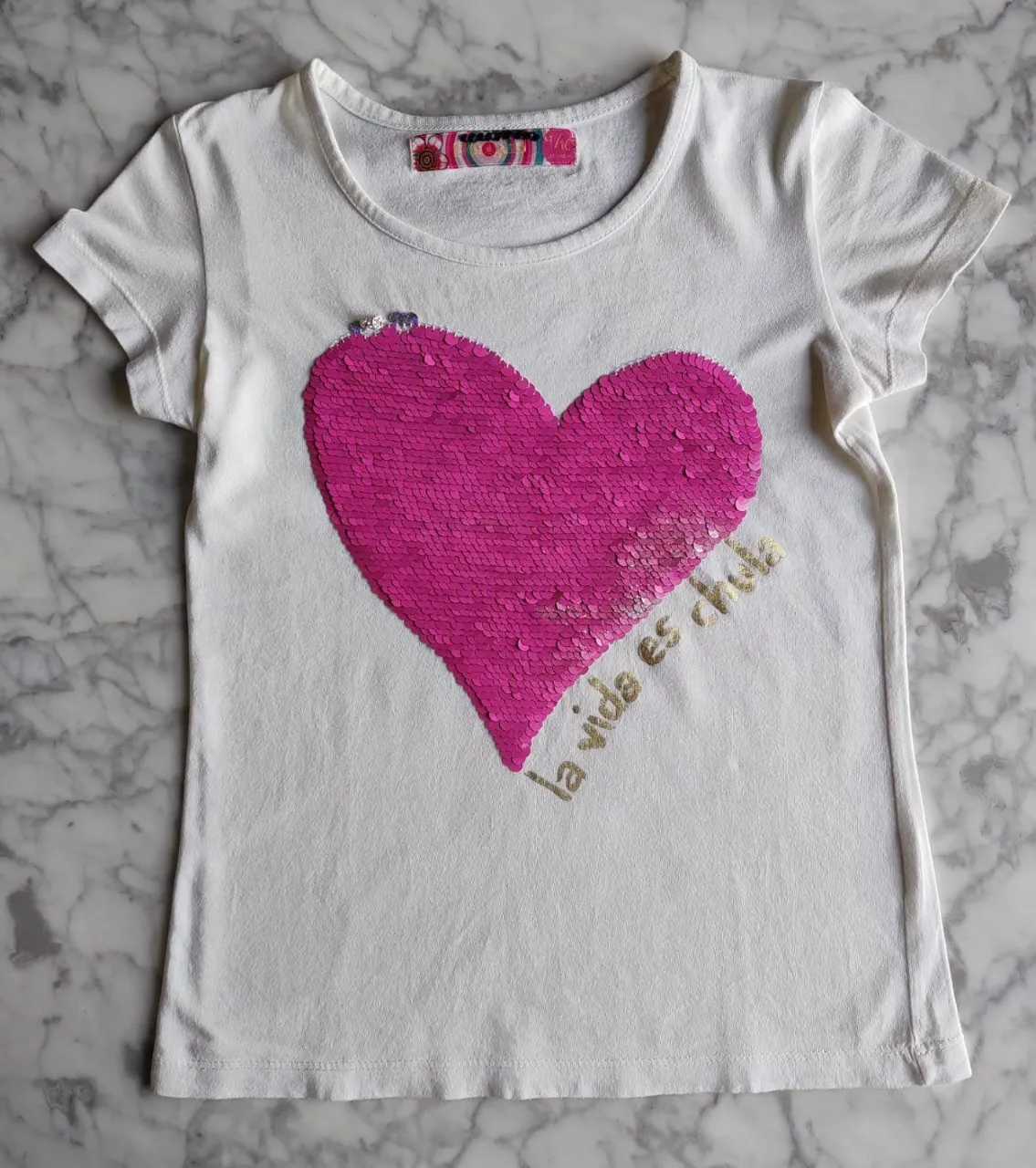 Remera blanca de algodón con un corazón de lentejuelas reversibles en color rosa y plateado. Tiene la frase 'la vida es chula' estampada en dorado. Ideal para un look casual y divertido.