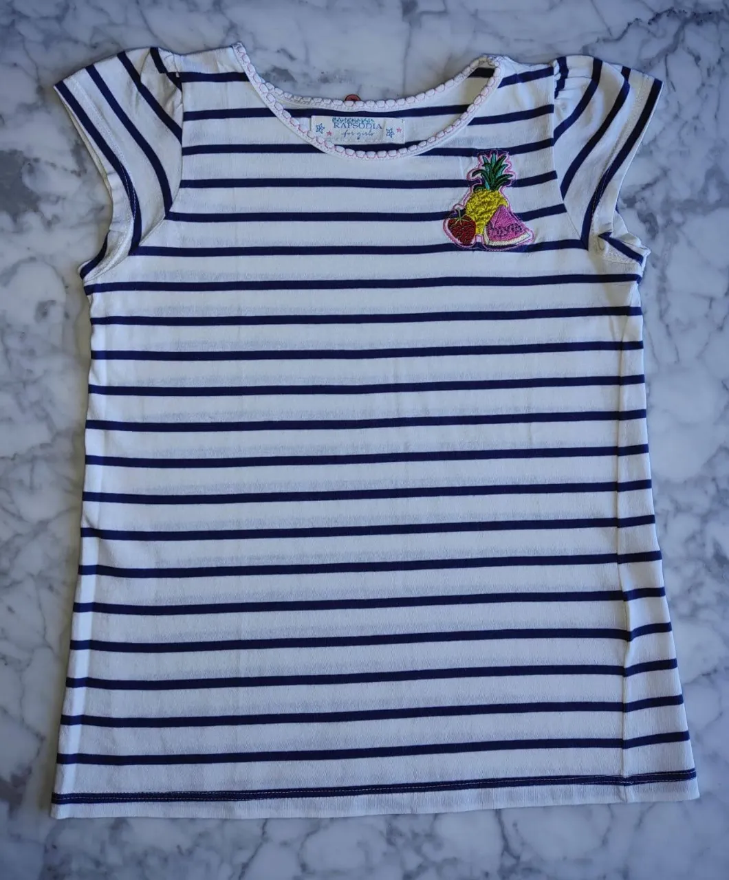 Remera de niña a rayas horizontales blancas y azules, con un aplique de frutas en el frente. Cuello redondo con detalle bordado y mangas cortas. Ideal para un look casual y fresco.