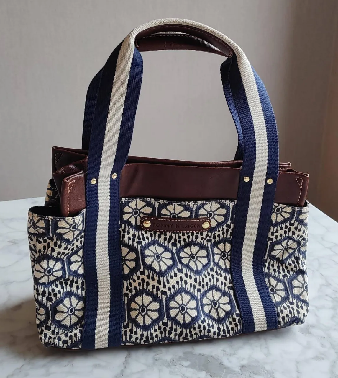 Cartera Tommy Hilfiger de mano, con estampado floral en tonos azul y blanco. Detalles en cuero marrón y manijas a rayas. Ideal para un look casual y moderno.
