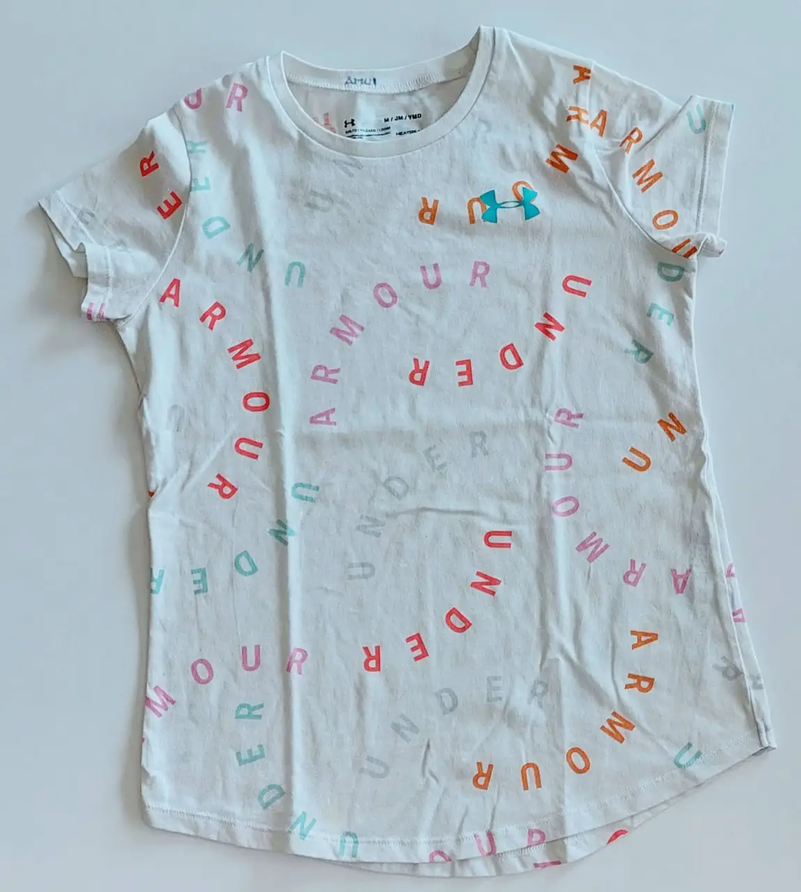 Remera deportiva blanca de la marca Under Armour para niña, con estampado de la marca en colores rosa, naranja y celeste. Cuello redondo y manga corta. Ideal para actividades físicas.