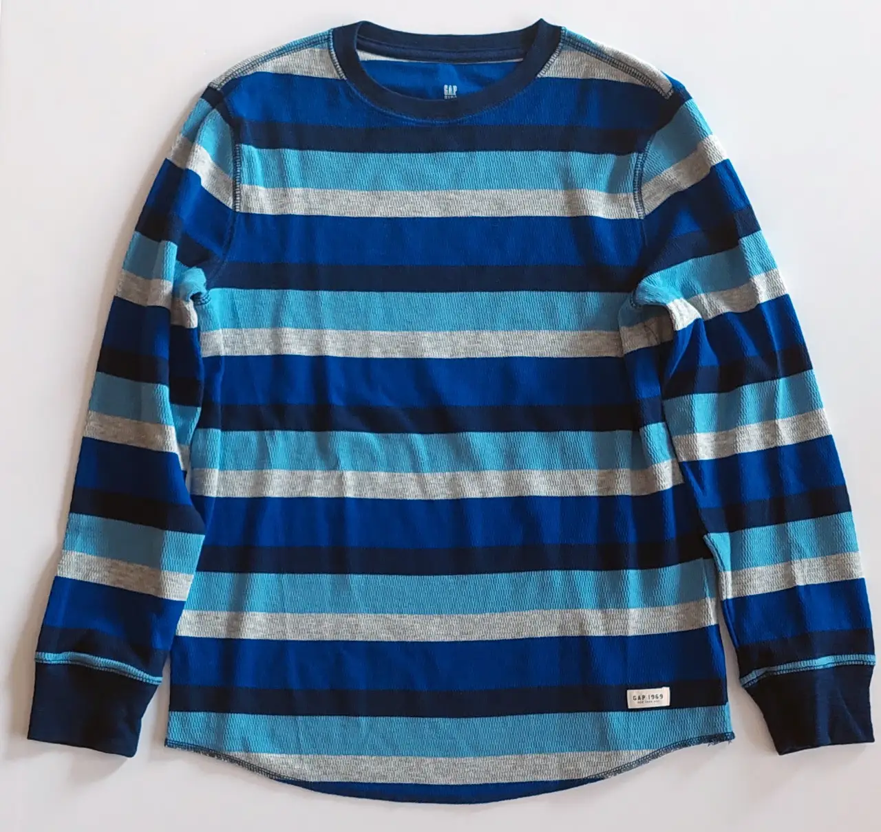 Remera de manga larga para niño, a rayas horizontales en tonos azules y grises. Con el logo de GAP en la parte inferior.