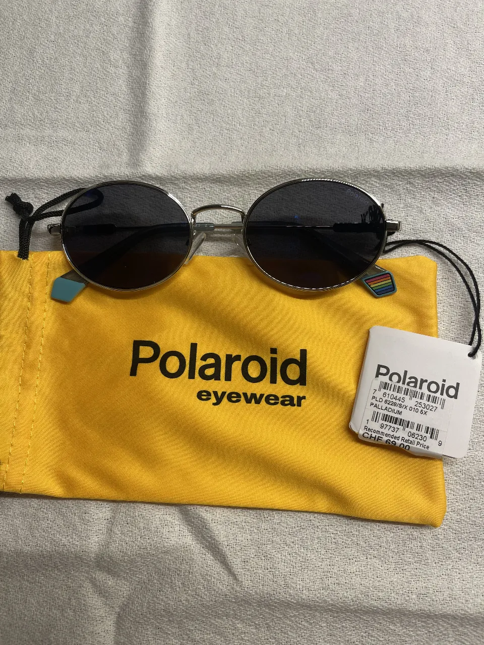 Anteojos de sol Polaroid, con armazón plateado y lentes oscuros. Incluye estuche amarillo con el logo de la marca y etiqueta con información del producto.