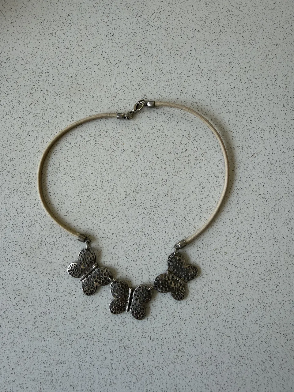 Collar delicado con tres mariposas plateadas texturizadas, ideal para darle un toque especial a tu look. Cierre metálico y cordón fino color beige.
