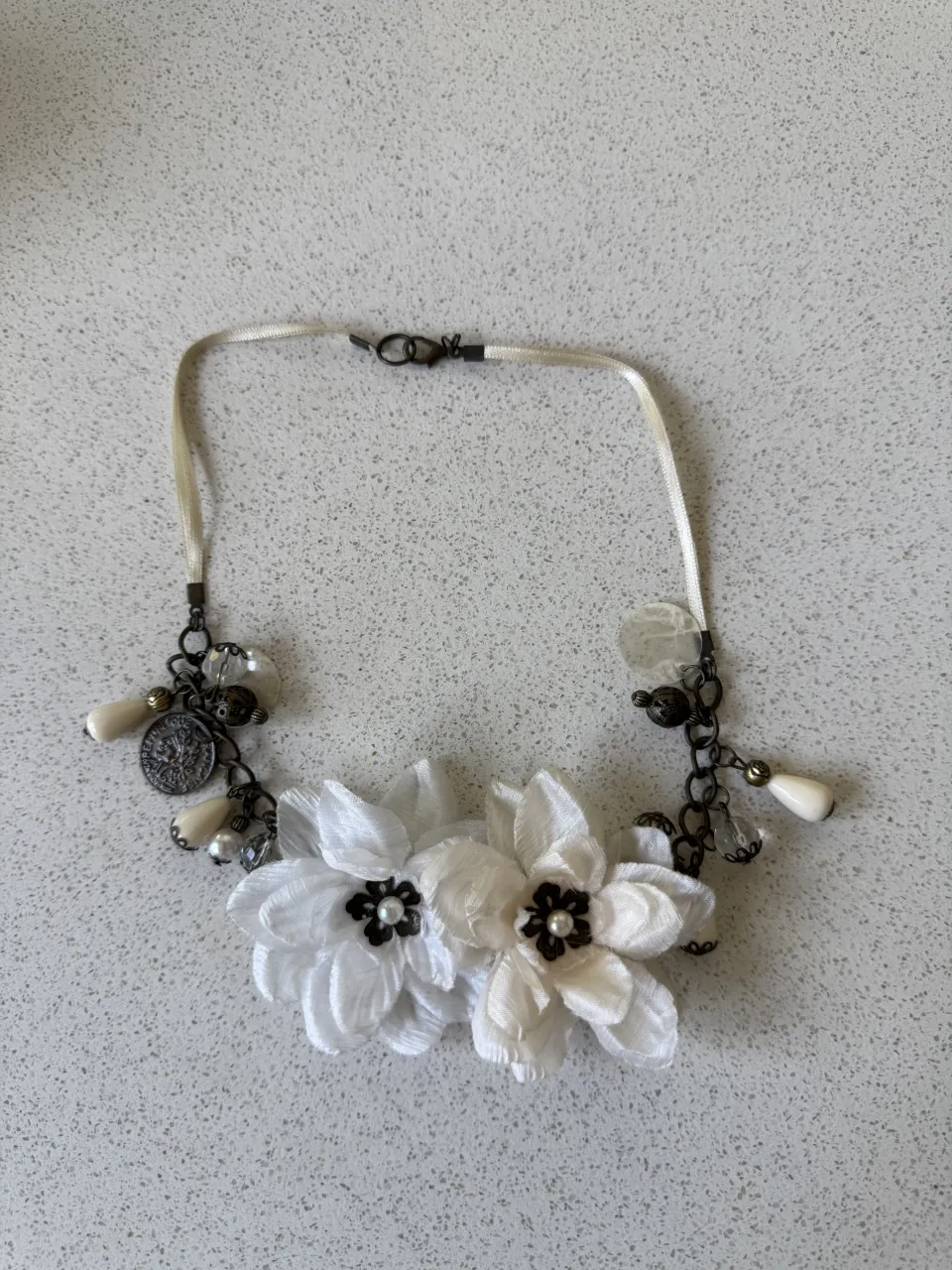 Collar delicado con dos flores blancas de tela y detalles colgantes como perlas y monedas antiguas. Un accesorio único para darle un toque romántico y vintage a tu look.