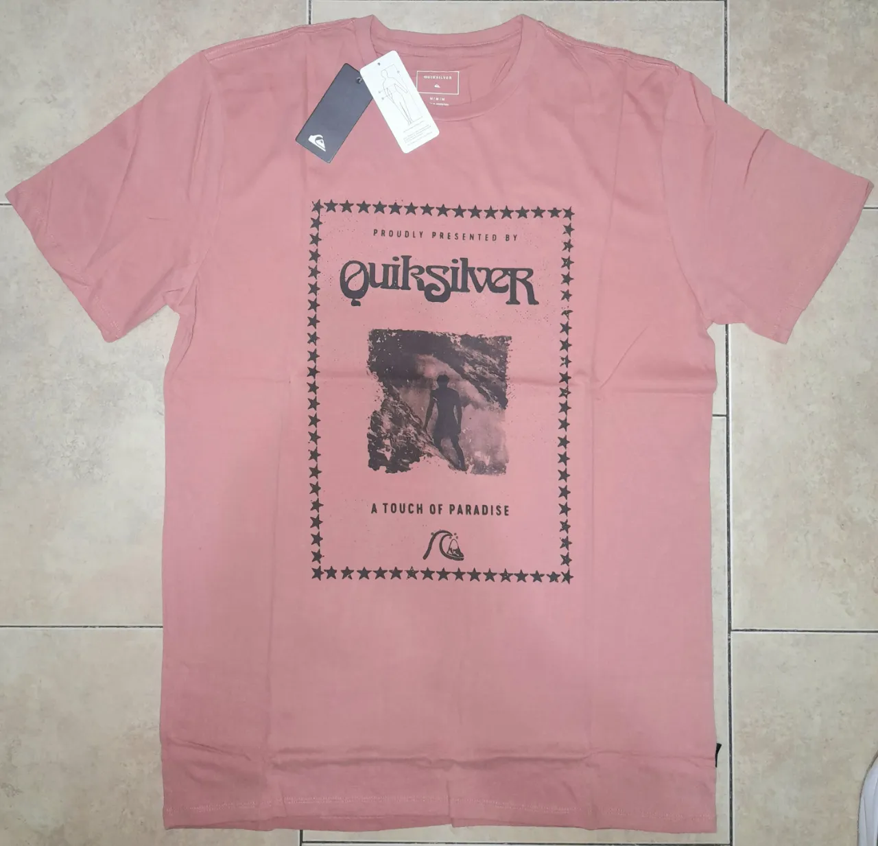 Remera manga corta de Quiksilver, color rosa pálido, con estampa "Proudly Presented By Quiksilver" y una imagen de un surfista. Ideal para un look casual y playero.