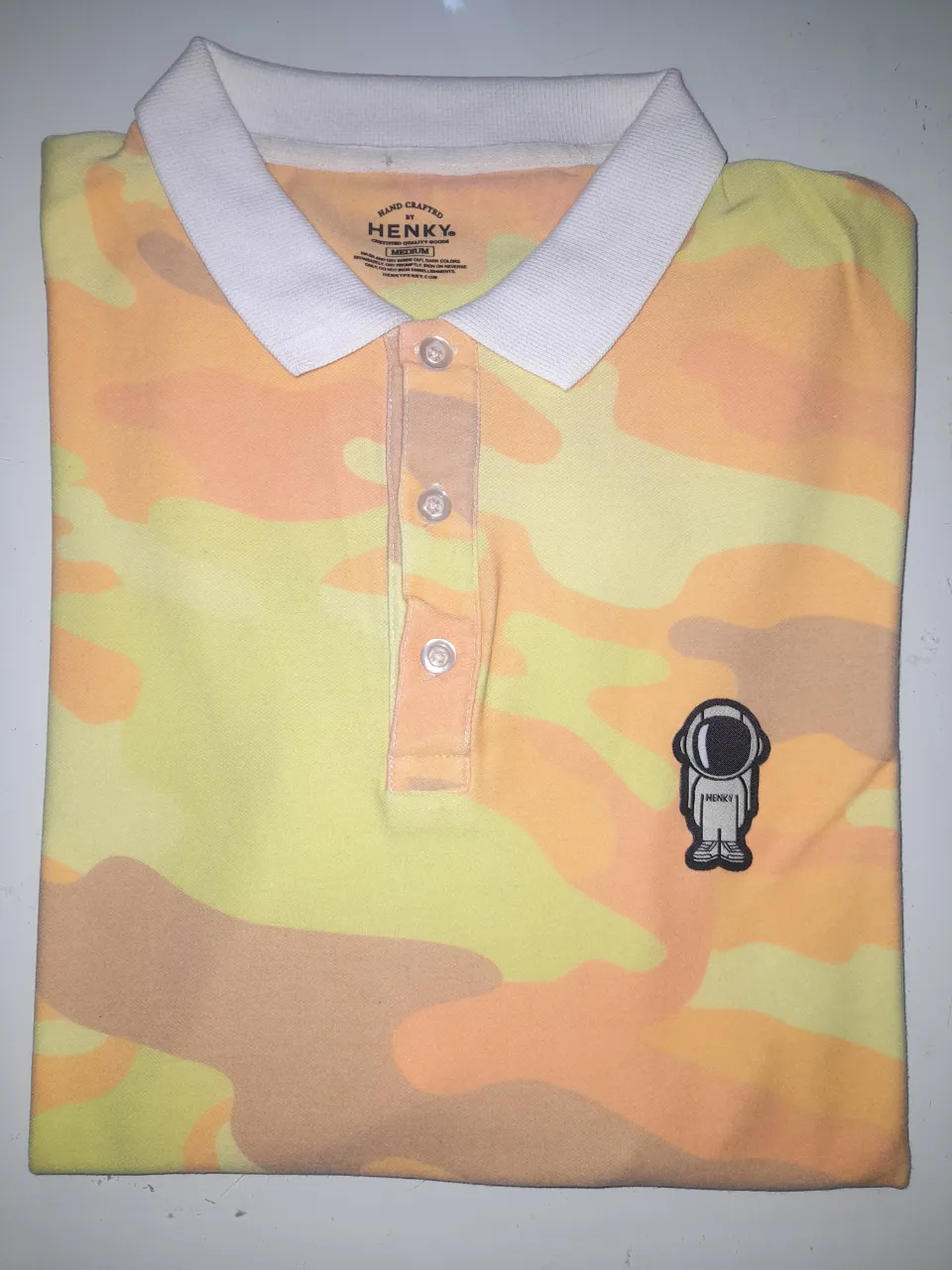 Remera polo Henky con estampado camuflado en tonos amarillos y naranjas. Diseño con astronauta bordado. Ideal para un look casual y moderno.