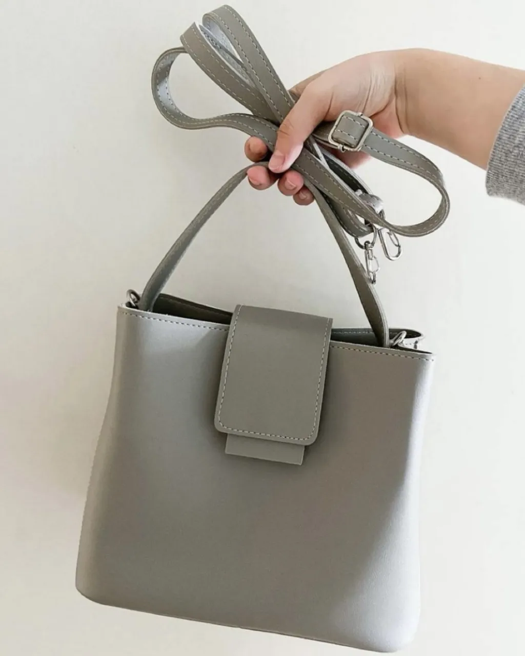 Bolso gris de cuero sintético, con correa regulable y manija corta. Cierre superior con solapa y broche. importada de Corea del sur