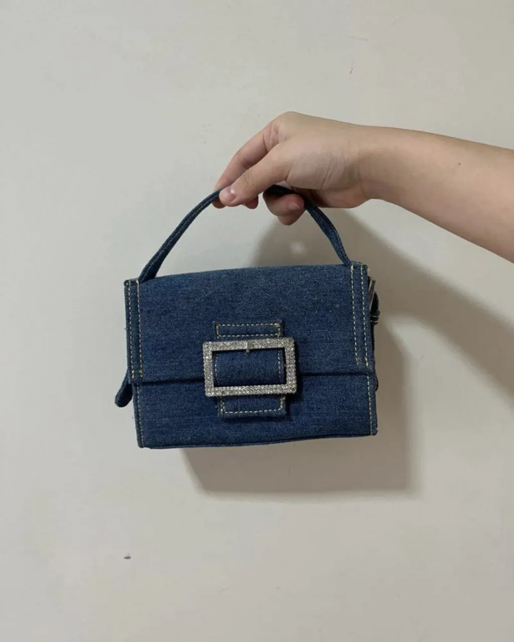 Cartera de jean azul con hebilla grande plateada con strass. Tiene correa larga regulable y manija corta. Ideal para un look canchero y moderno.