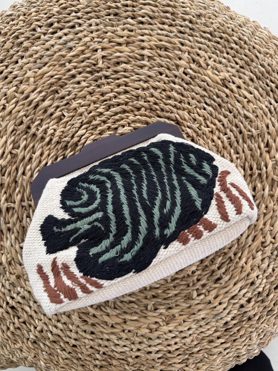 Cartera de mano con bordado de diseño abstracto en tonos verde, negro y marrón sobre base cruda. Posee un cierre de madera oscuro.